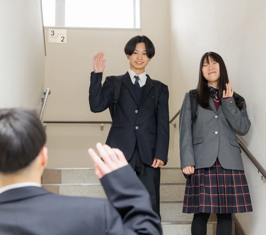 校舎内の階段で、学生同士が笑顔で手を振り合い挨拶を交わしている学校生活の一場面