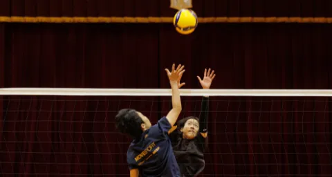 ネット越しに、生徒がジャンプしてボールに手を伸ばしているバレーボールのプレーの様子
