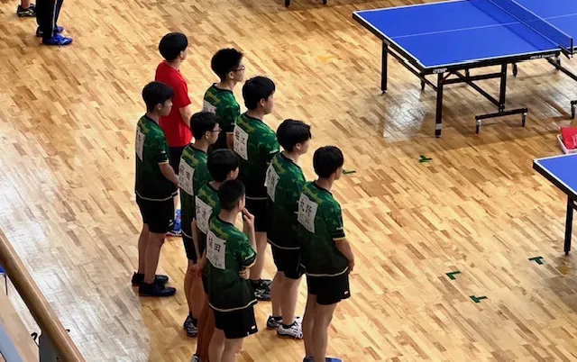 卓球台のそばで、緑色のユニフォーム姿の部員が整列している様子